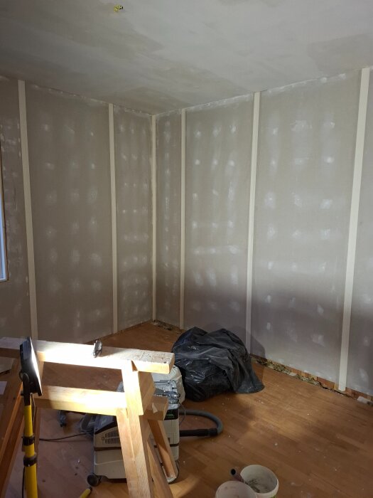 Renovering av rum med gipsade väggar och tak, verktyg och byggmaterial synliga på golvet och ett arbetsbock framme i en hörna.