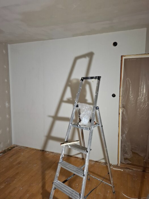 Stege mot gipsad vägg i pågående rumsrenovering med plast över dörröppningen och trägolv.
