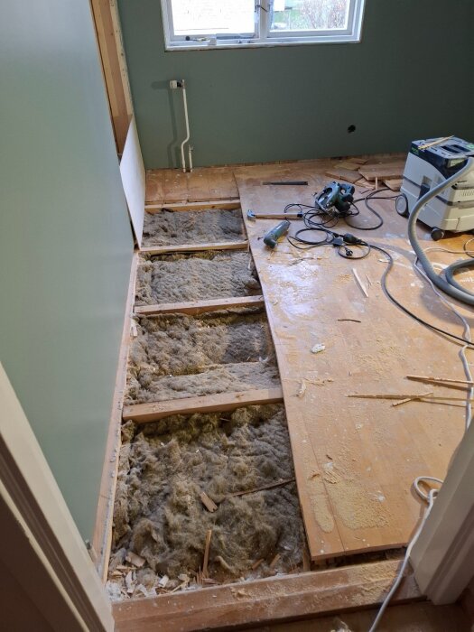 Renovering av ett rum med borttagna parkettbrädor och synlig isolering, en såg och dammsugare står på golvet.