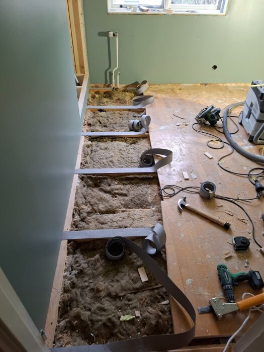 Renovering av rumsgolv där gamla parkettgolvet tagits bort. Synlig isolering och verktyg såsom en hammare, måttband och elverktyg ligger på golvet.