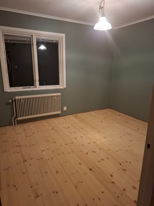 Rum med trägolv, grönmålade väggar och taklampa. Ett fönster med element under är synligt.