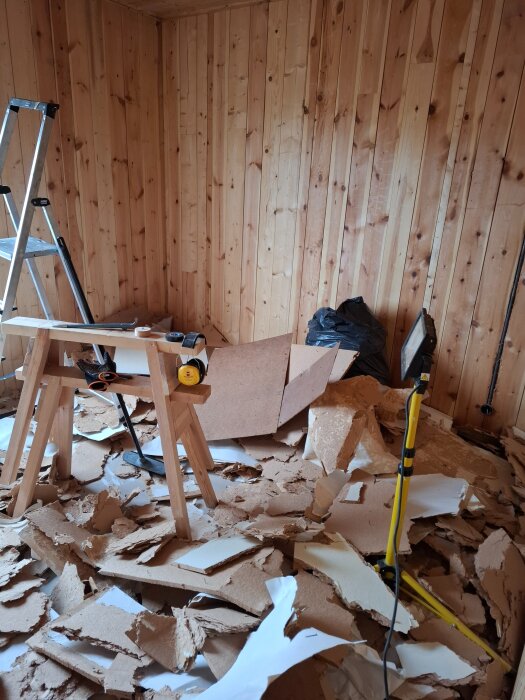 Rum under renovering med träpaneler, stege och arbetsbord; golvet täckt av rivet pappersmaterial och byggskräp, omgivna av byggverktyg.