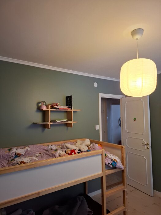 Vardagsrum med grön vägg, våningssäng och två hyllor ovanför. En lampa i taket och dörr i bakgrunden.