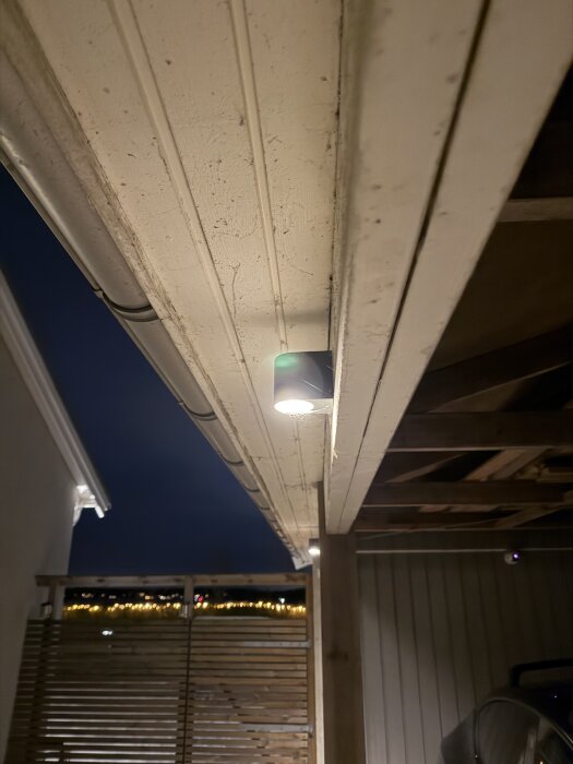 Carportens takstol och belysning i kvällsljus, med en del av husets fasad och skymtande staket i bakgrunden.