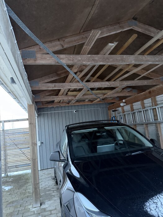 Carport med synligt träbjälklag och en svart bil parkerad under taket. Ljusgrå träpanel på väggarna. Snö syns på bilen och marken.