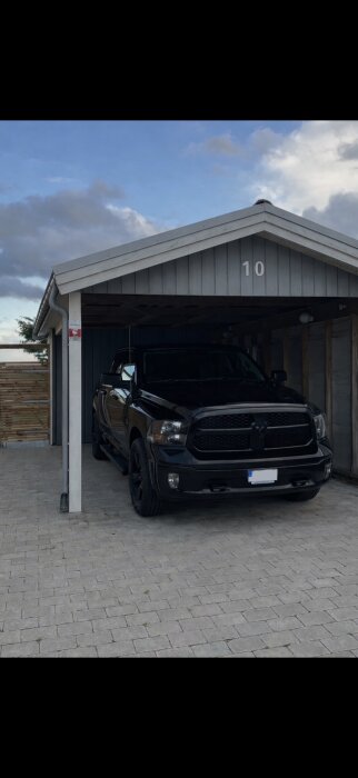 Carport med grå fasad, nummer 10, och en svart bil parkerad under. Ränna syns till vänster.
