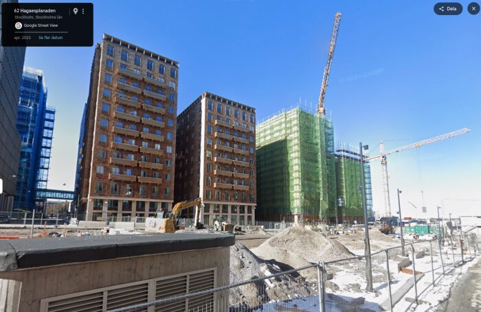 Byggarbetsplats med höga hus och byggkranar i Stockholm, inkluderar ett täckt hus och en grävmaskin i förgrunden.