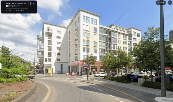 Gata i Stockholm med moderna flerbostadshus, träd, och parkerade bilar under en blå himmel.