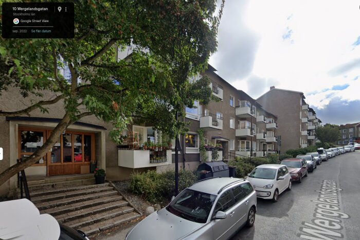 Flerfamiljshus med balkonger och träd i förgrunden i en förort i Stockholm. Parkerade bilar längs gatan.