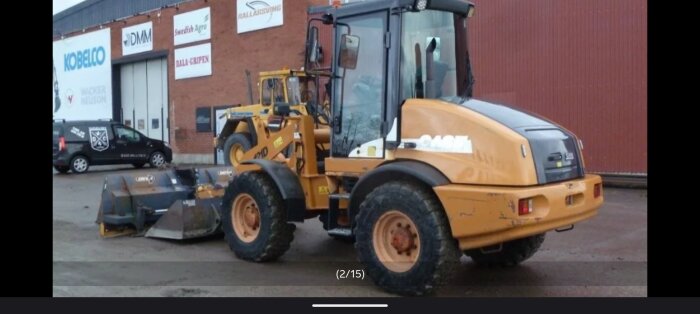 Case 121D hjullastare parkerad vid industribyggnad med skyltdäck på väggen bakom och en annan lastare i bakgrunden.