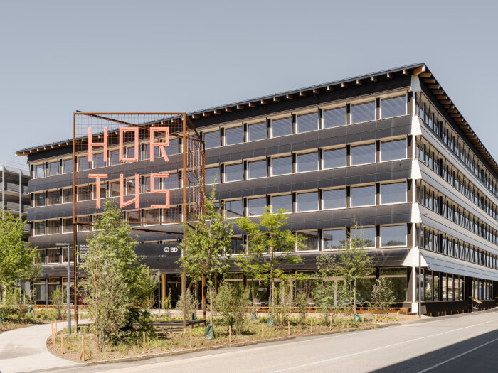 Kontorsbyggnaden "Hortus" med modernistisk arkitektur och en stor metallstruktur med texten "Hortus" framför, omgiven av träd.