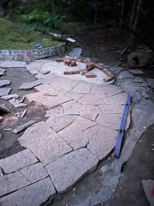 Byggande av uteplats med oregelbundna stenplattor, några tegelstenar och en vattenpass. Omringad av grönska och gångväg.