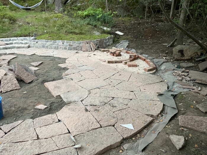 Stenlagd uteplats under konstruktion med ojämnt formade stenar och synliga fogar. Bruk och tegelstenar runt kanterna, naturmiljö i bakgrunden.