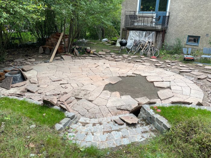 Stenläggning på en cirkelformad uteplats i en trädgård, omgiven av gräs och ett hus med balkong i bakgrunden.
