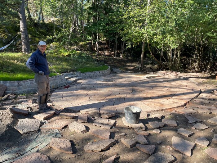 Man står vid en ofärdig uteplats av stora, oregelbundna stenar på en solig dag, omgiven av träd och en mur. En hink syns i förgrunden.