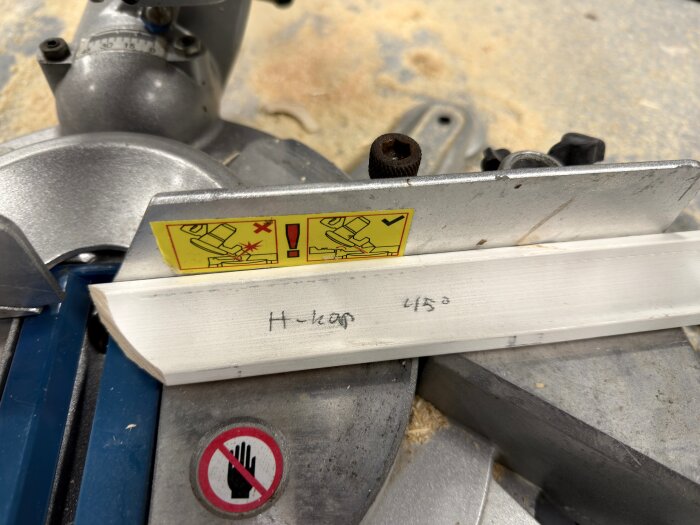 Vinklat trästycke på geringssåg med varningsskyltar. Handskriven text "45°" på listen. Frågetecken om rätt mätpunkt, övre eller undre kant av listen.