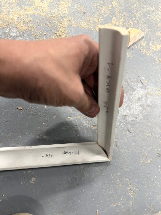 Hand håller ihop två träbitar skurna i 45 graders vinkel för att bilda ett hörn, markerade med siffrorna 1 och 2, på ett dammigt golv.