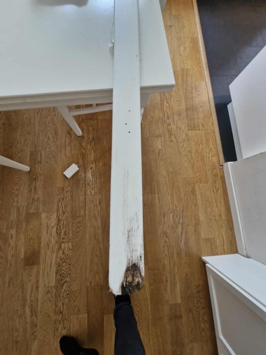 Vitt dörrfoder med rötangrepp i nedre delen ligger på ett parkettgolv bredvid ett vitt bord.