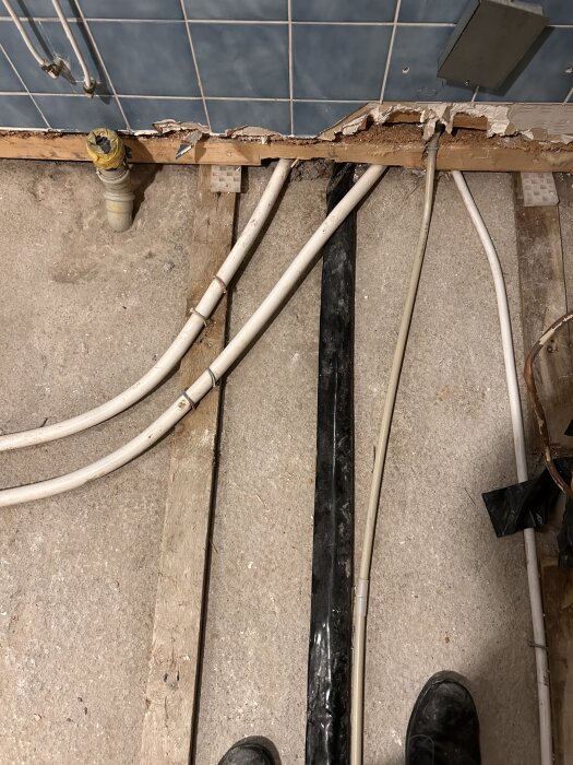 Svarta isoleringsskyddet kring kallvattenrör i 70-tals hallrenovering, sett ovanifrån med synliga plaströr och trasigt trä längs väggen.