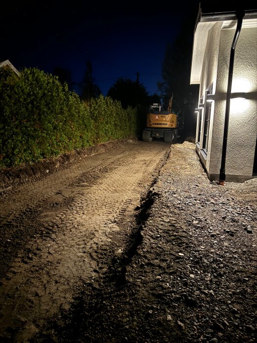 Uppfart under natten med grävd mark och en Liebherr-grävmaskin i bakgrunden, belyst av en byggnads utomhusbelysning.