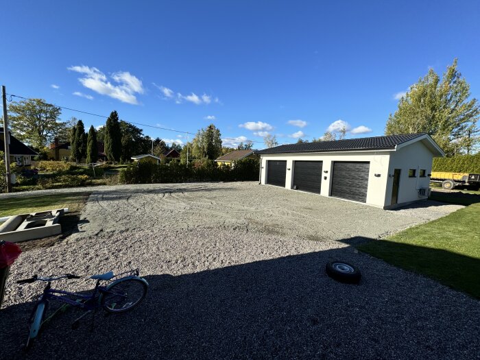 Uppfart under arbete med ny markbeläggning, visar en grusplan framför ett garage med flera portar, omgiven av grönska och en cykel i förgrunden.