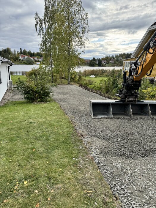 Grävmaskin i arbete vid en uppfart, omgivande gräsyta, och sjö i bakgrunden under en molnig himmel.