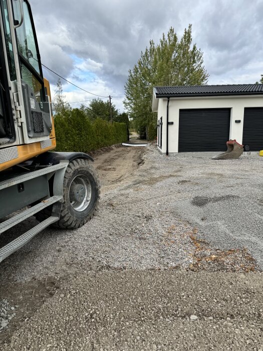 Grävmaskin vid urgrävd uppfart med lager av grus och fiberduk, vy mot garage och träd i bakgrunden.