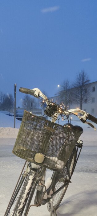 Cykel med snöig korg och julbelysning står på vintrigt trottoar med snö och lägenhetshus i bakgrunden under skymning.
