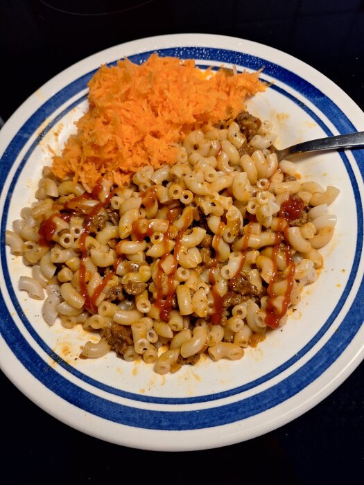 Pasta med köttfärs och ketchup samt riven morot på en tallrik med blå kant, med en sked vid sidan.