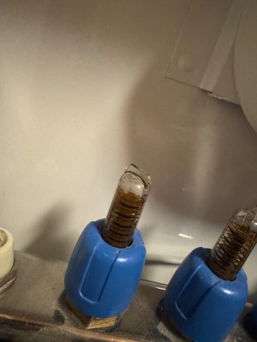 Blåa kopplingar med glasrör fyllda med brun vätska, möjligen sand, på ett rörsystem. Indikerar problem med ett värmesystem.