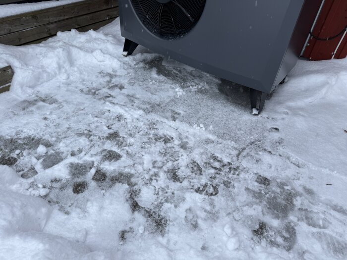 Is och snö under en värmepump med synligt fläktelement, beläget nära en trävägg, på en frusen markyta.
