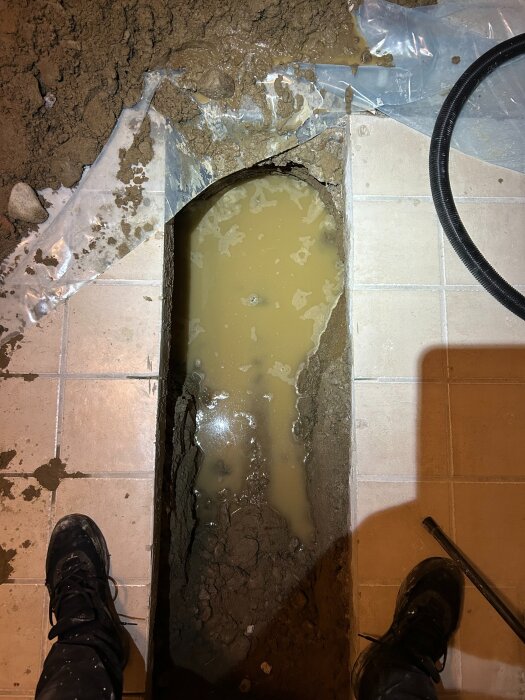 Grävd grav i källargolv med vattenansamling, omgiven av jord och plastduk, del av avloppsbyte.