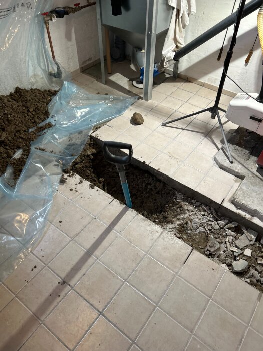 Spade stående i ett delvis uppgrävt hål på ett kakelgolv i en källare, omgiven av jord och utrustning.