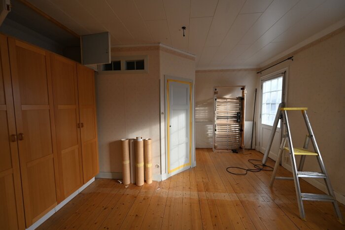 Rum under renovering, med träplankgolv, garderober, en stege, rullar av papper och ett inbyggt badrum från 90-talet borttaget.