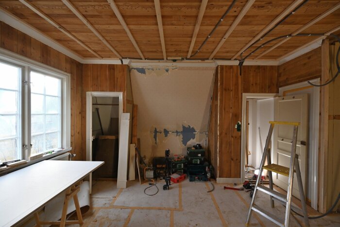 Rum under renovering med vederväggar, verktyg på golvet, en stege och ljust fönster. Vägg innehåller borttagen tapet. Två dörröppningar syns.