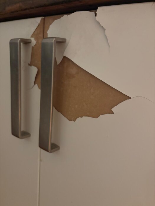 Spruckna och avflagade vitlackerade köksluckor med två moderna metallhandtag, skadad ytbeläggning synlig.