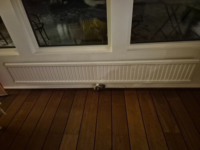Vit radiator monterad under ett fönster på ett mörkt trägolv.