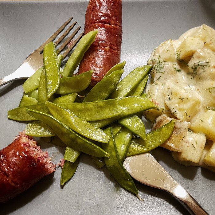 Grön sockerärtor, korv och potatis i gräddsås på en tallrik med gaffel och kniv.