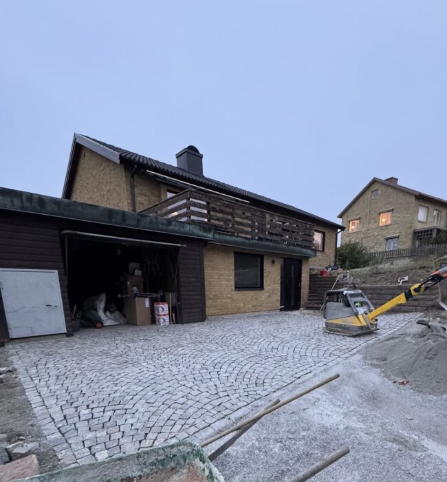 Stenbelagd uppfart framför ett gult tegelhus, omgiven av verktyg och byggmaterial.