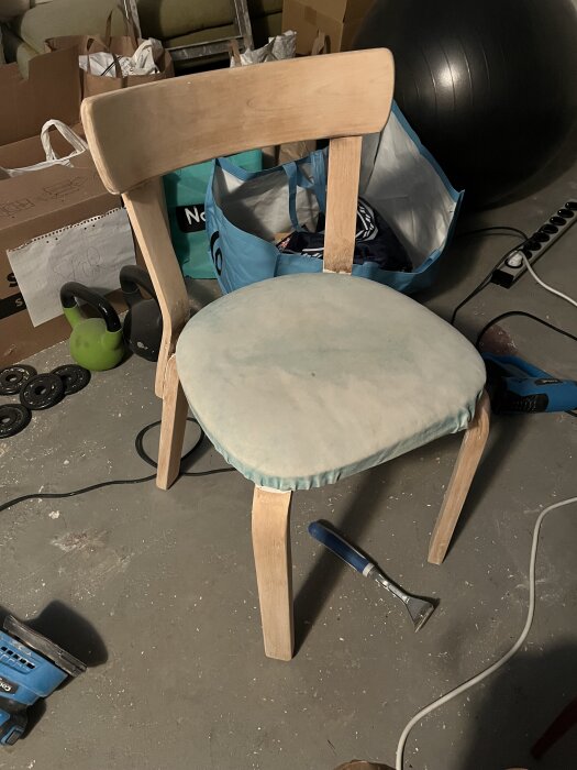 Trästol under renovering med avskrapad färg, omklädd sits, värmelampa och slipverktyg synliga på betonggolv omgivna av verktyg och förvaringslådor.