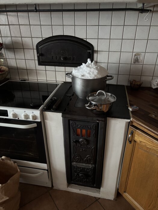Gjutjärnsspis med en stor gryta fylld med snö på spisplattan, en mindre kastrull bredvid, i ett kök med vita kakel.