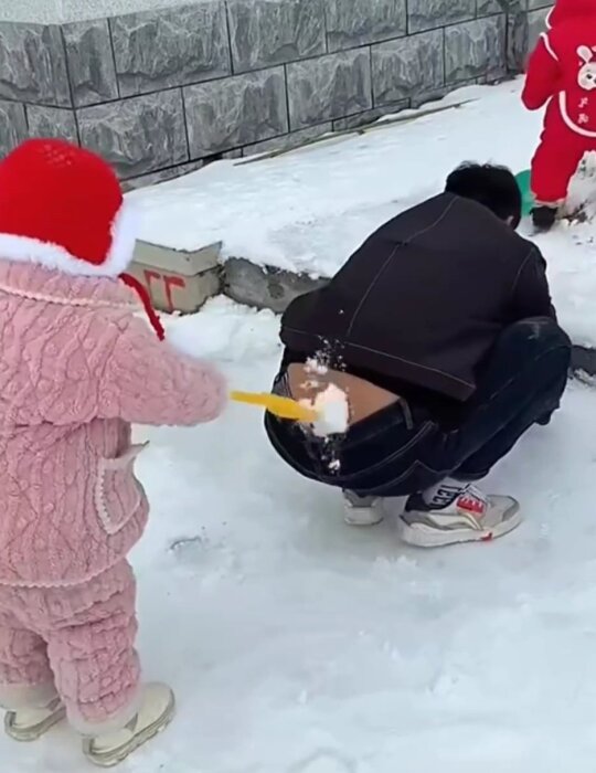 Litet barn i rosa overall leker i snön och placerar en snöboll på en hukande persons bak, bredvid en byggnad med stenbeklädnad.