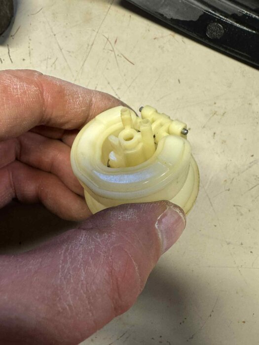 Hand håller en liten, ljus plastdel med flera cylindriska utsprång, mot en ljus bakgrund.