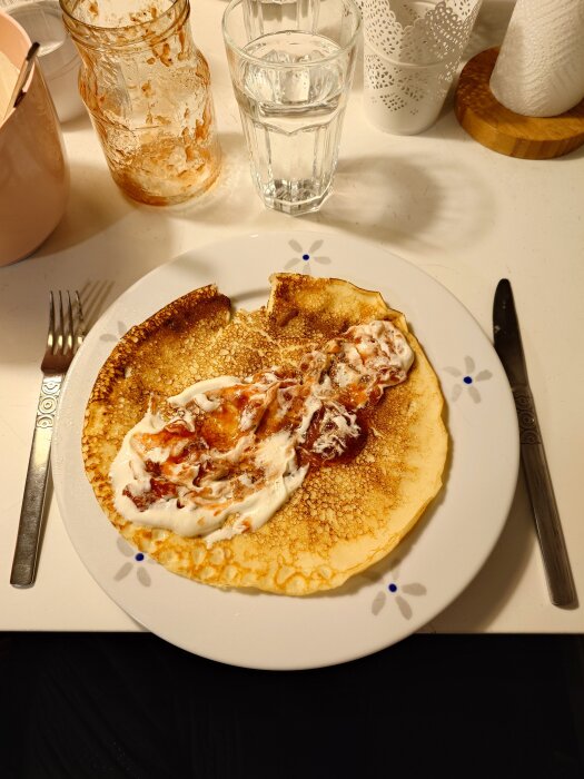Pannkaka med sylt och grädde på en tallrik, omgiven av bestick och ett glas vatten på ett bord.