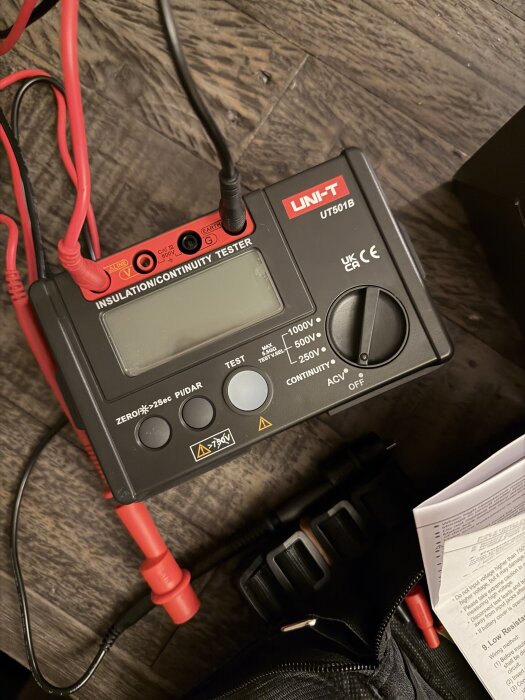 Digital isolations- och kontinuitetstestare med märkningen UNI-T UT501B, kablar anslutna, ligger på ett träbord bredvid en manual och verktygsväska.