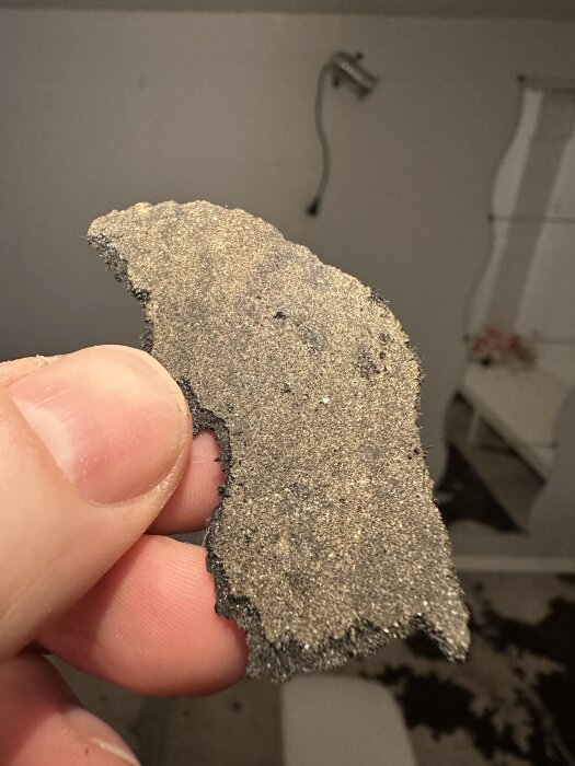Närbild av en hand som håller en bit av gammalt svart material med dammig yta, potentiellt lim eller matta från 50-talshusets källargolv.