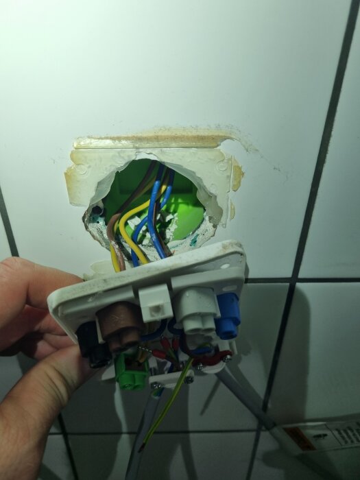Elinstallation i väggen med synliga kablar och uttag, där en hand håller i en lös strömbrytare framför kakelplattor.