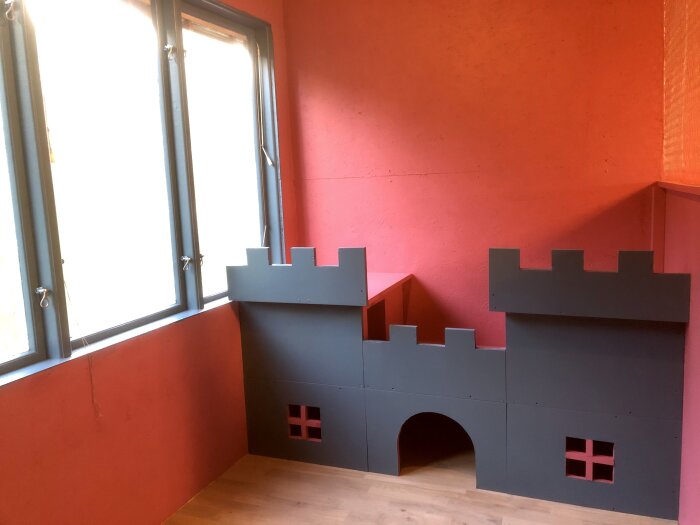 Kaninhus i form av ett litet slott med torn och fönster, placeras i ett rum med röda väggar och trägolv.