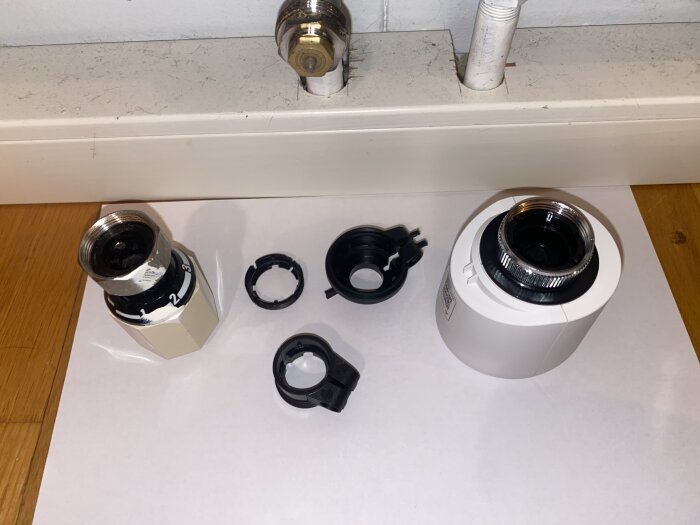 Termostat och adaptrar placerade på ett vitt papper framför ett elementrör på ett trägolv.