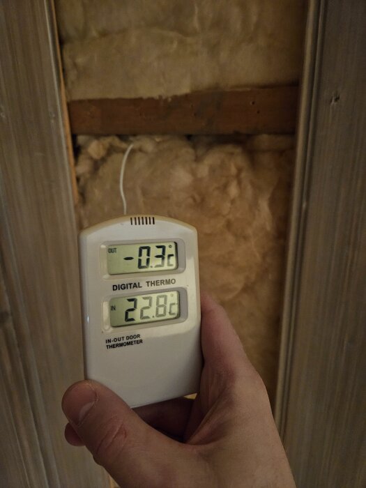 Digital termometer visar temperaturändring från -0,3°C utanför till 22,8°C inuti, framför isoleringsmaterial och trävägg.